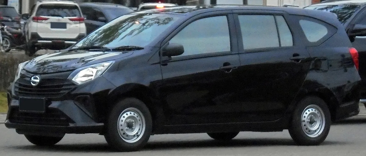 Daihatsu Sigra hitam tampak depan samping dengan velg baja, di jalan dengan mobil lain di latar belakang