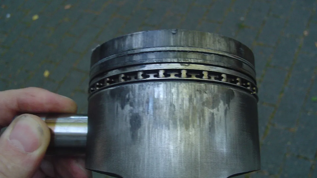 Piston logam yang dipegang tangan, dengan noda gelap dan goresan memanjang di badan sampingnya serta ring piston yang kotor