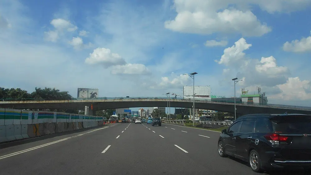 Jalan akses Bandara Soekarno-Hatta di Kota Tangerang dengan jembatan layang, langit berawan, dan mobil keluarga hitam di lajur kanan