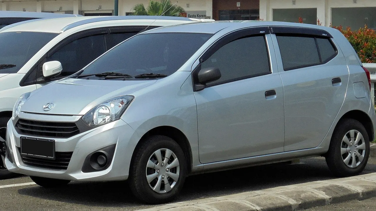 Daihatsu Ayla generasi pertama tipe 1.0 D+ berwarna perak terparkir di tepi jalan, terlihat lampu utama besar, gril sempit, dan velg baja dengan dop