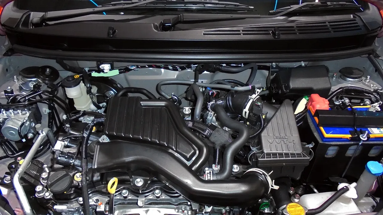 Ruang mesin Daihatsu Ayla generasi pertama tipe 1.2 R Deluxe dengan penutup mesin hitam, saluran udara masuk, tangki cairan pendingin, dan aki di sisi kanan