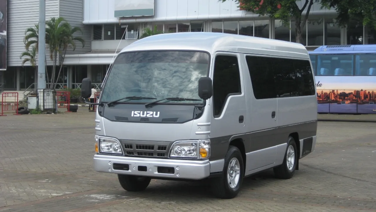 Isuzu Elf NKR55 microbus berwarna perak dengan atap tinggi, terparkir di pelataran beraspal Jakarta, tampak depan samping kiri