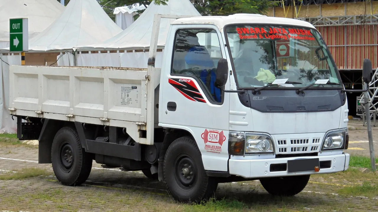 Isuzu Elf NHR 55 berkabin putih dengan bak besi model jungkit, terparkir di halaman berumput dekat tenda putih
