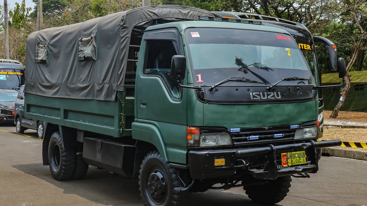 Truk Isuzu Elf NPS 75 berwarna hijau tentara dengan bak bertutup terpal, ban off-road, dan bemper depan besi, terparkir di Monas Jakarta