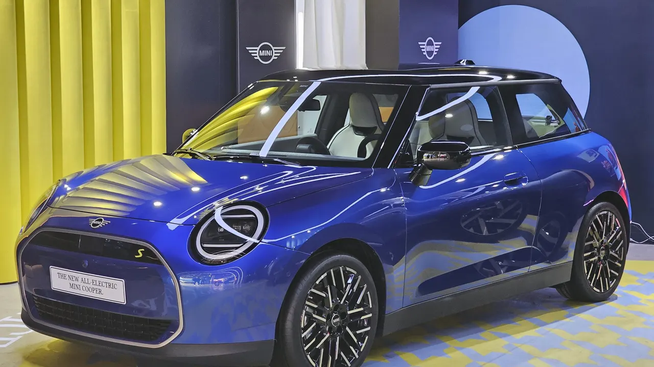 MINI Cooper SE listrik generasi J01 berwarna biru dengan atap hitam dan lampu bulat berbingkai, dipajang di ruang pamer berlatar kuning