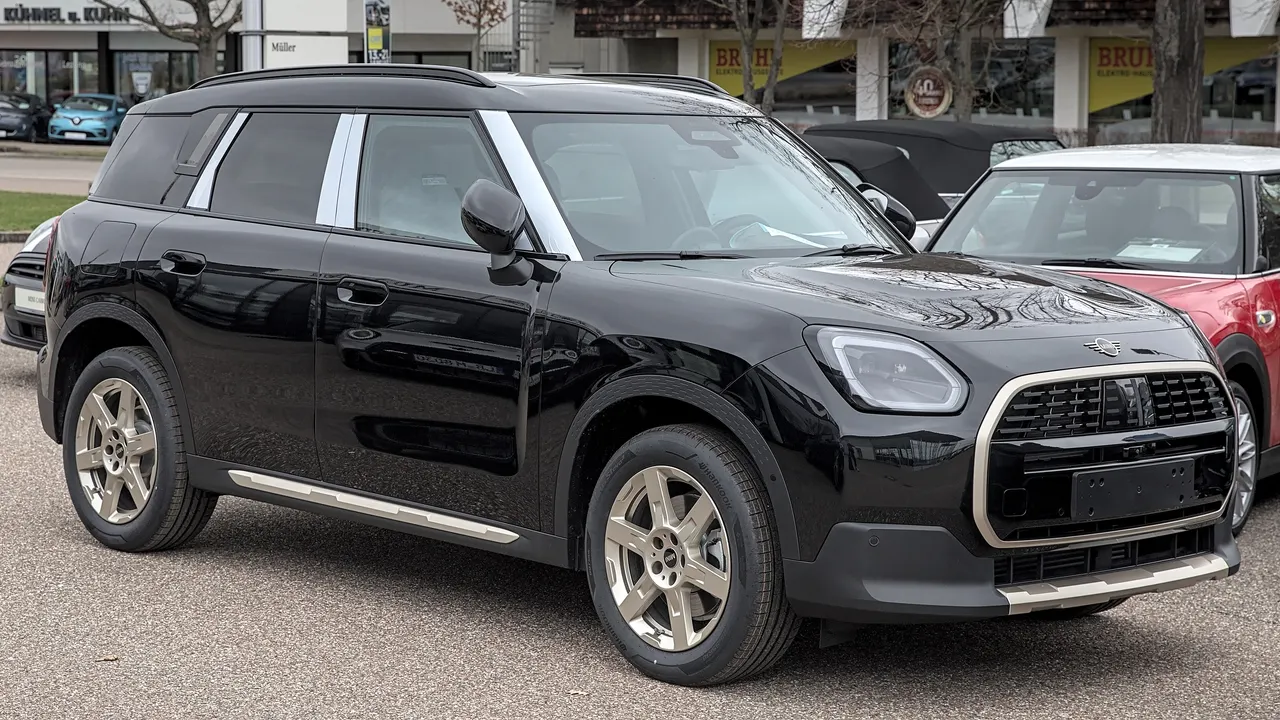 MINI Countryman generasi U25 berwarna hitam dengan pilar putih, gril segi delapan berbingkai perak, dan pelek dua warna, terparkir di area parkir terbuka