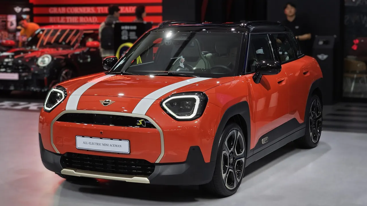 MINI Aceman SE berwarna merah oranye dengan atap hitam dan dua garis putih di kap mesin, dipajang di panggung pameran mobil