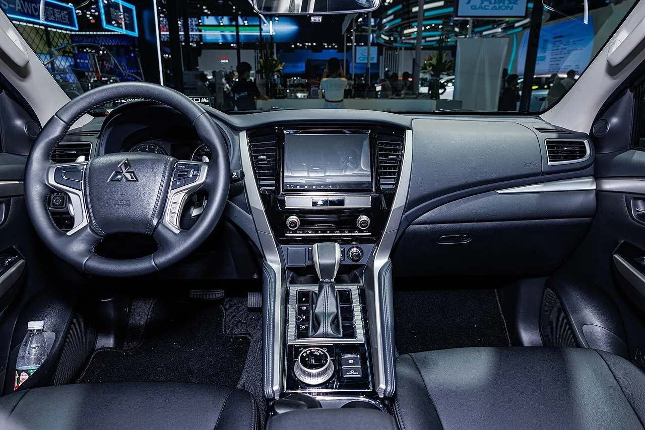 Dasbor dan konsol tengah Mitsubishi Pajero Sport dengan tuas transmisi otomatis, kenop putar pemilih mode di bawahnya, dan tombol rem parkir elektronik