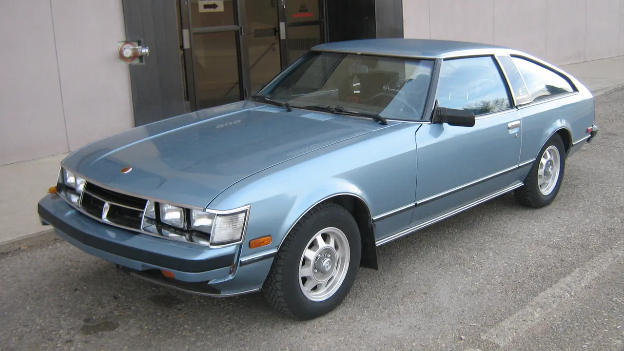 Toyota Celica Supra generasi pertama berwarna biru muda metalik terparkir di pinggir jalan, terlihat empat lampu utama kotak dan bodi liftback panjang