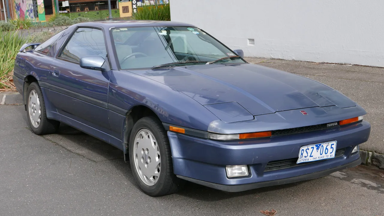 Toyota Supra MA70 tahun 1986 berwarna biru keunguan dengan lampu utama pop-up tertutup, terparkir di tepi jalan Melbourne