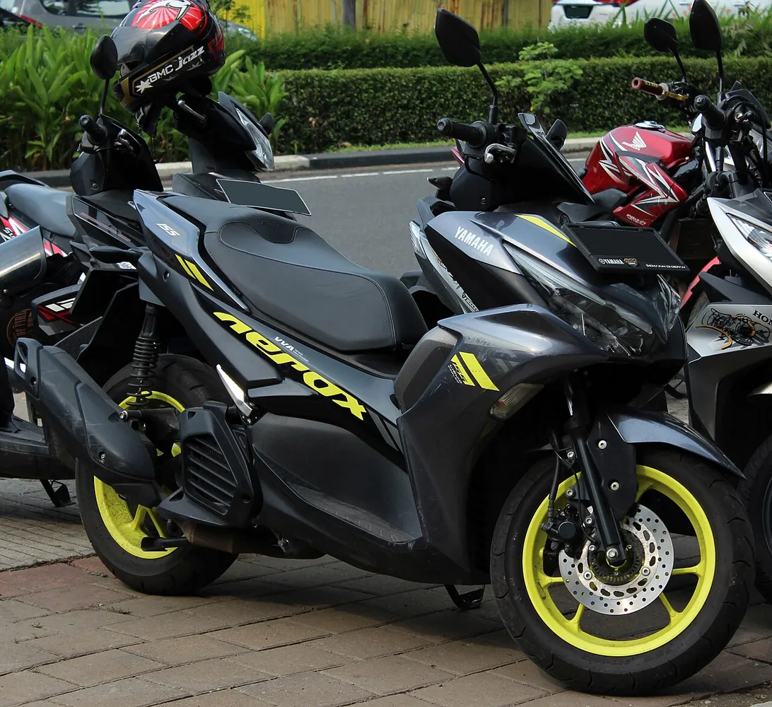 Yamaha Aerox 155 VVA abu-abu dengan pelek kuning terparkir di trotoar di antara sepeda motor lain