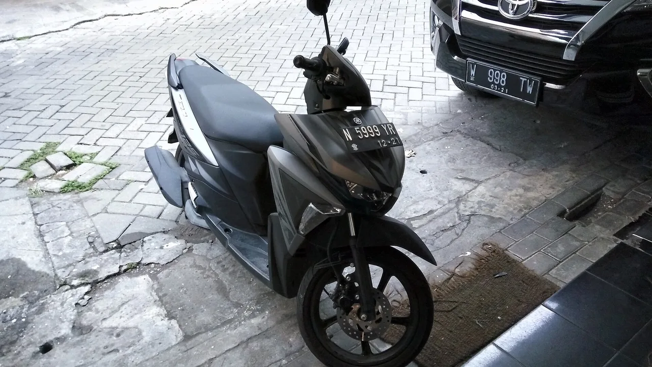 Yamaha Soul GT abu-abu gelap dan hitam terparkir miring di halaman berpaving, dengan sebuah SUV hitam di belakang kanannya