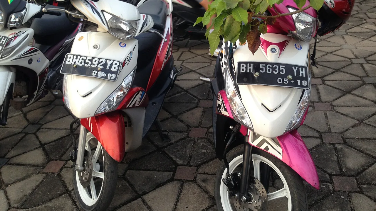 Bagian depan dua unit Yamaha Mio J, satu merah putih dan satu merah muda putih, terparkir berdampingan di lantai paving