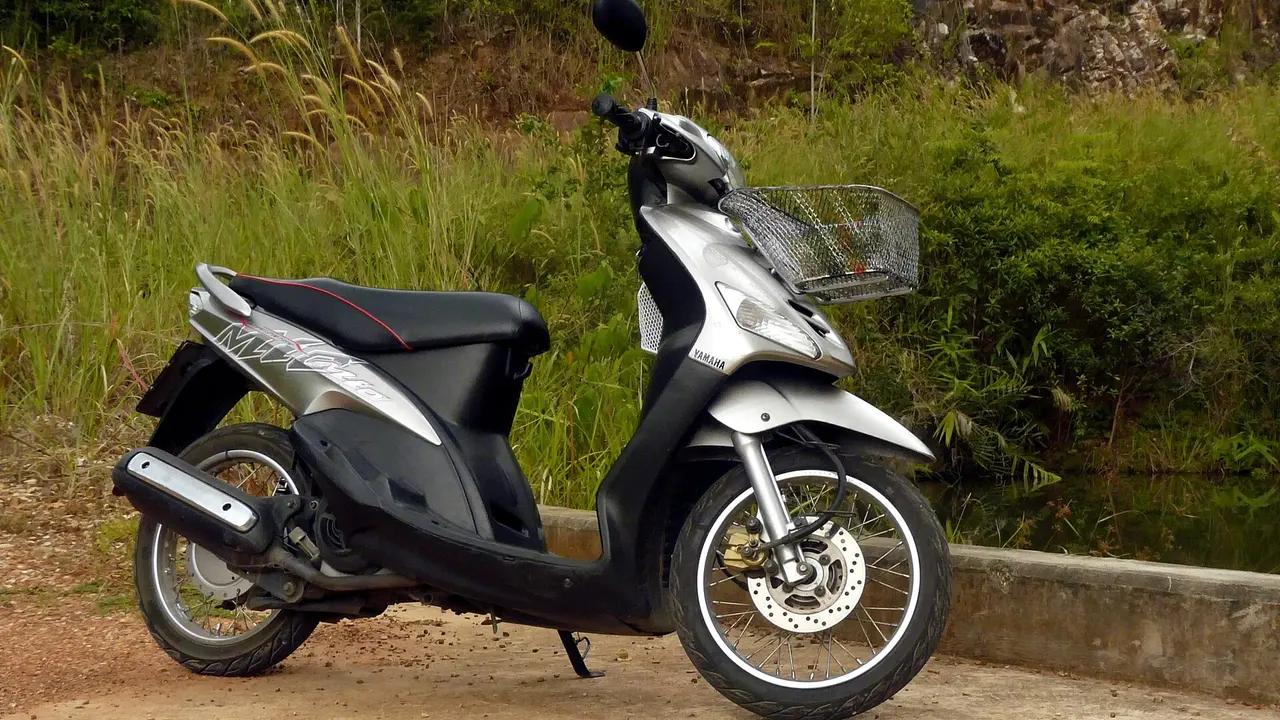Yamaha Mio generasi lama berwarna perak dan hitam dengan pelek jari-jari, rem cakram depan, dan keranjang besi di bagian depan, terparkir di pinggir jalan berumput