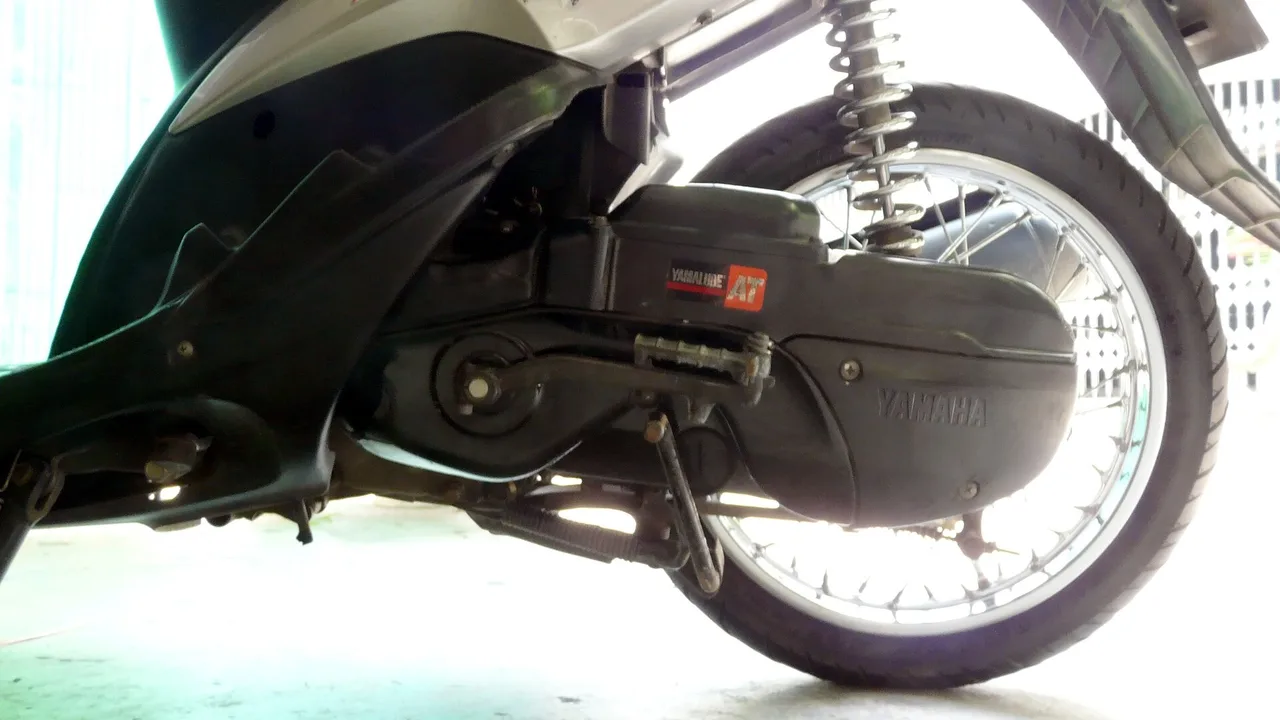 Bagian bawah skuter Yamaha Mio dari samping, memperlihatkan rumah CVT hitam bertulisan Yamaha, suspensi belakang tunggal, dan roda belakang berpelek jari-jari