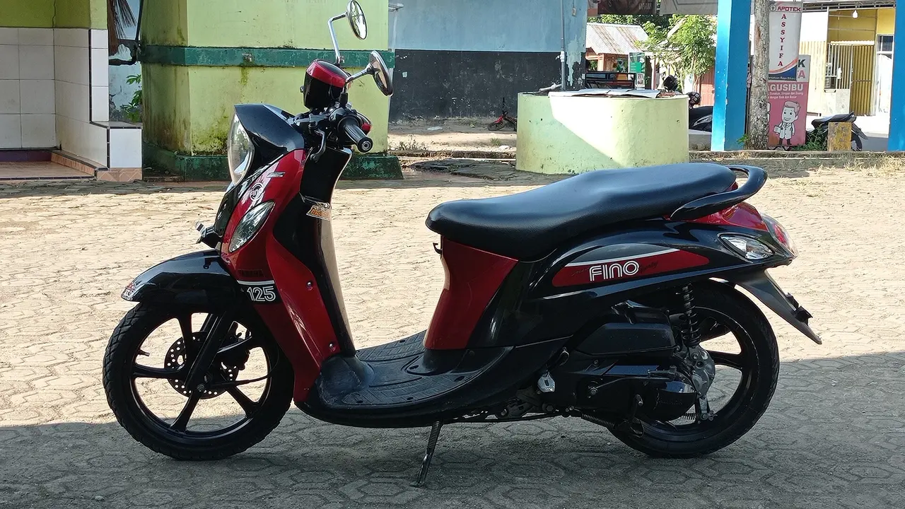 Yamaha Fino 125 merah hitam dengan tulisan Fino dan 125 di bodi, terparkir menyamping di halaman berpaving