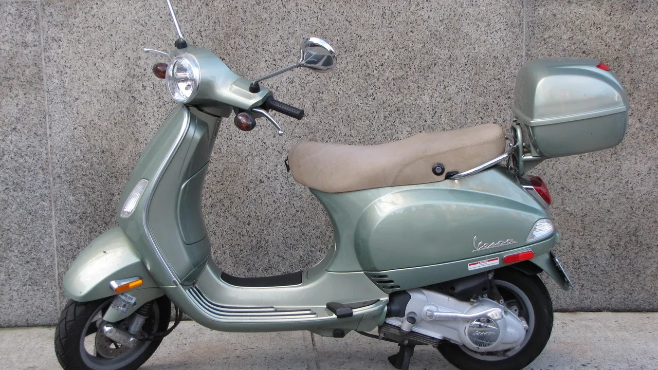 Vespa LX berwarna hijau keabu-abuan dengan jok krem dan kotak bagasi belakang, terparkir di depan dinding granit