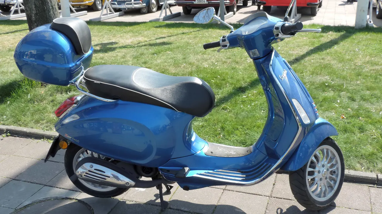 Vespa Sprint berwarna biru dengan jok hitam, velg alloy, dan kotak bagasi biru, diparkir di trotoar dekat rumput