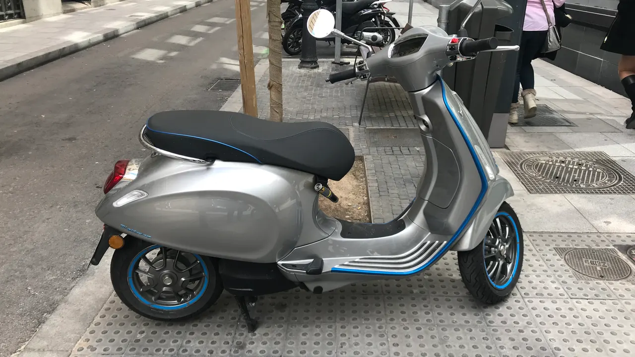 Vespa Elettrica berwarna perak dengan garis biru di bodi dan velg, diparkir di trotoar kota