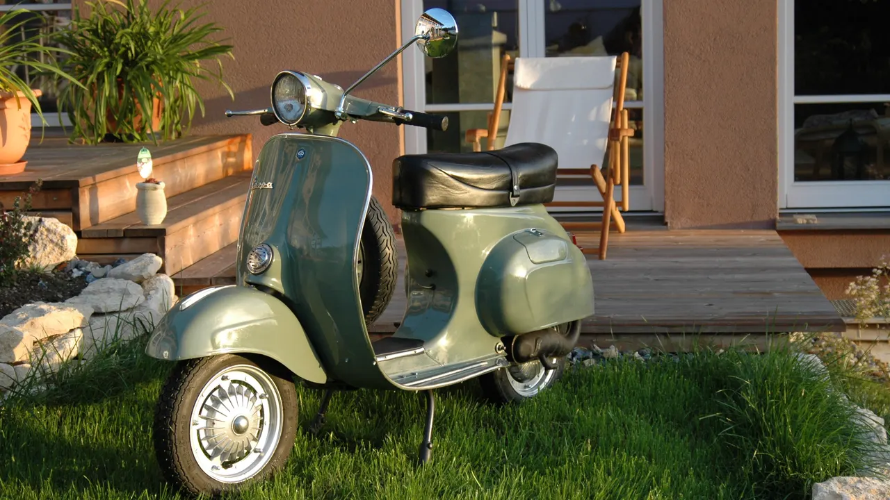 Vespa 125 Primavera ET3 klasik berwarna hijau keabu-abuan dengan jok hitam, ban serep di balik dek kaki, dan lampu depan bulat di setang, terparkir di halaman rumput