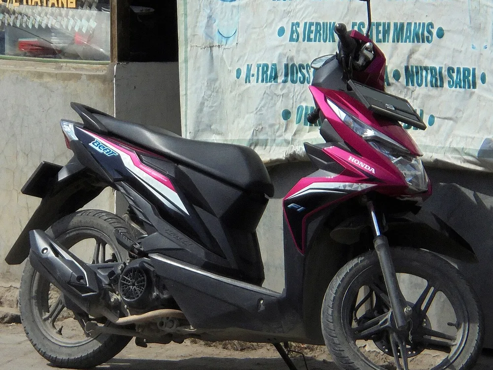 Honda BeAT eSP CBS-ISS warna merah muda dan hitam terparkir di depan warung, sisi kiri memperlihatkan blok mesin, CVT, dan knalpot