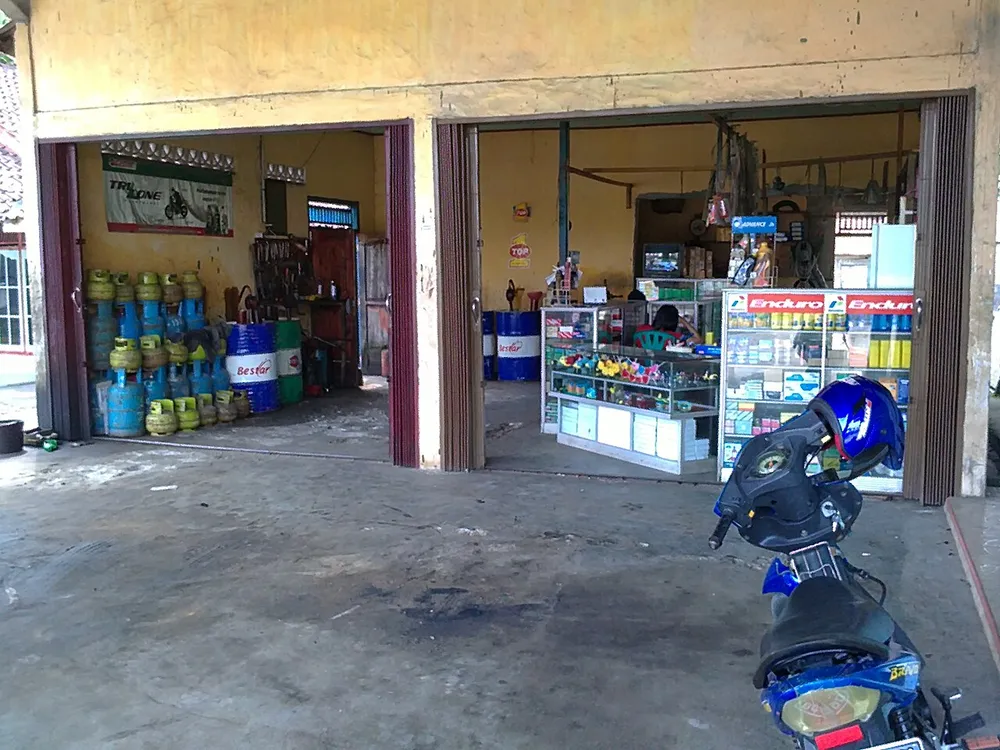 Bagian depan bengkel ganti oli kecil dengan rak botol oli, drum oli, tabung gas, dan sepeda motor terparkir